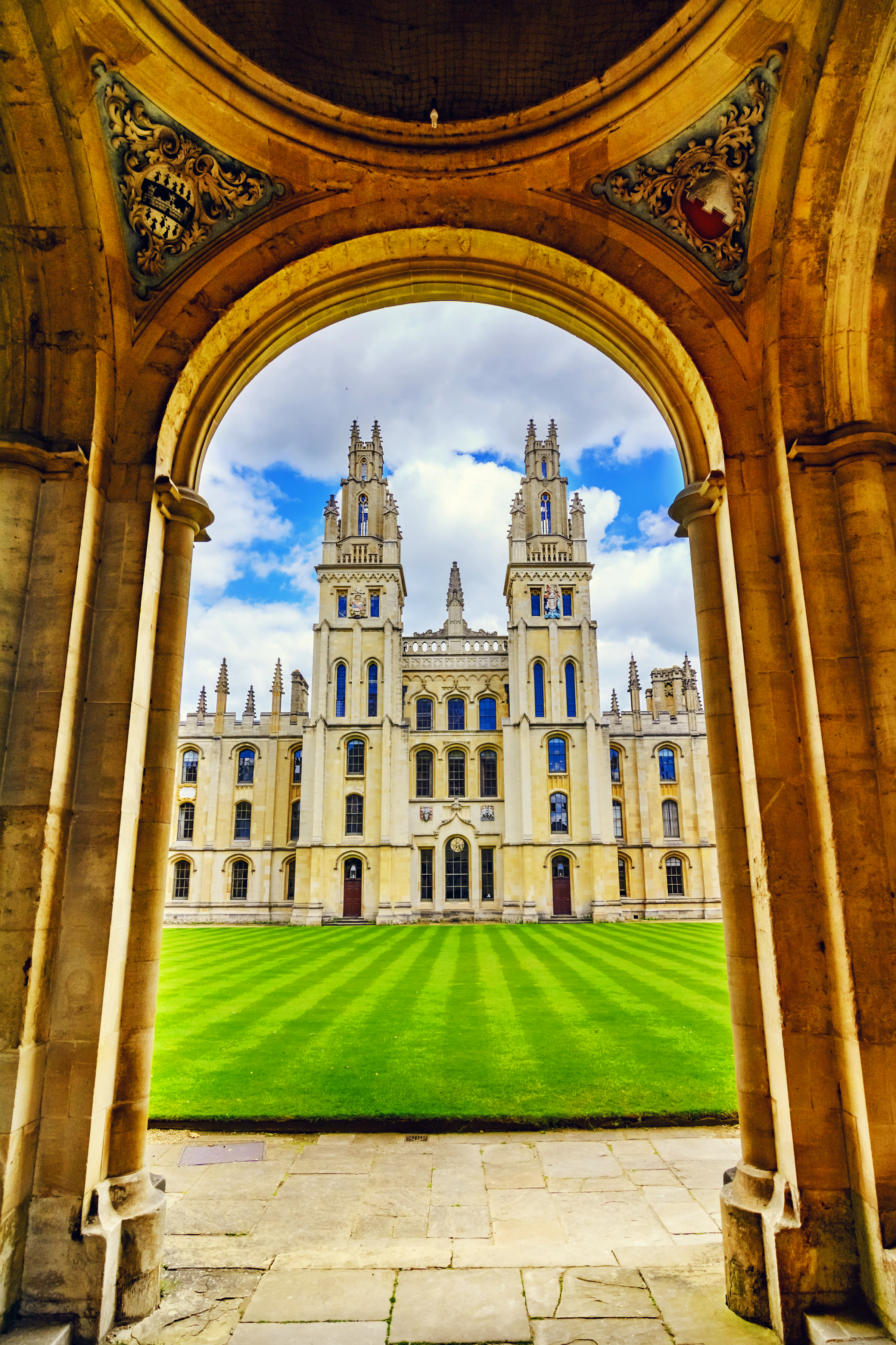 The twin gothic towers of All Souls College, Oxford, framed through a stone archway above a striped quad lawn