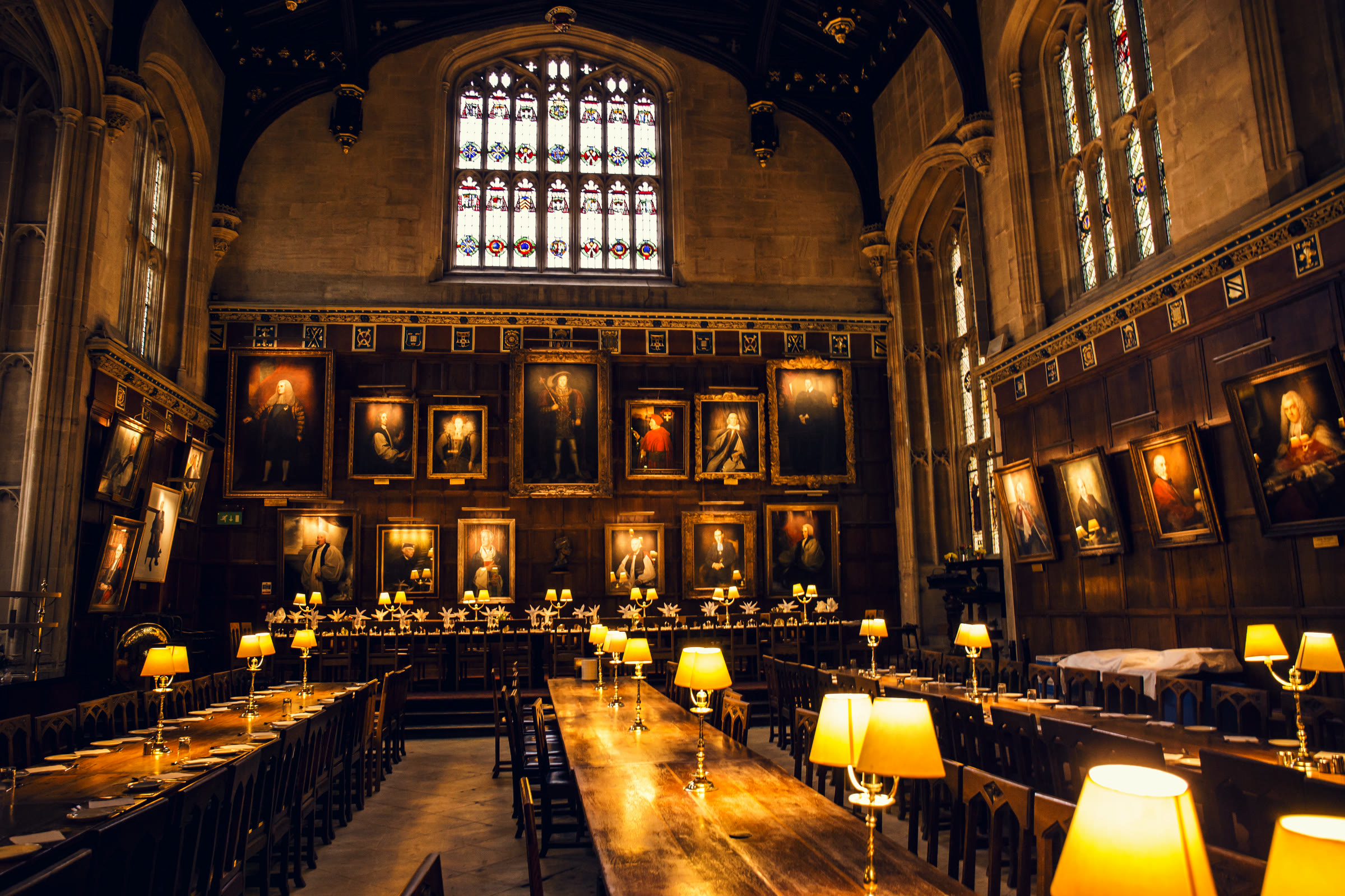 The lamplit dining hall of an Oxford college, with long oak tables, glowing brass lamps and portrait-lined panelled walls